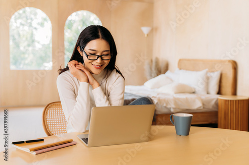Young teenage Asian woman wears sweater and glasses with pretty smile in living room. Happy and stylish girl using laptop with notebook and coffee. Winter portrait fashion lifestyle. Copy space © twinsterphoto