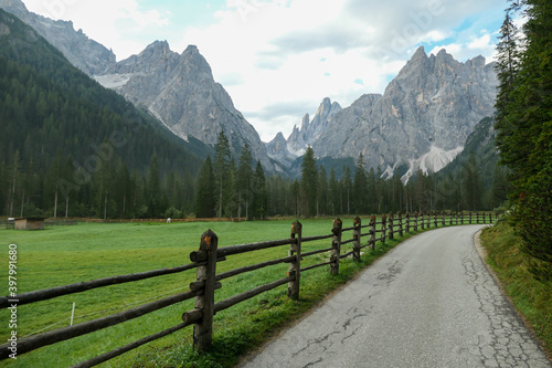 A gravelled road leading along a lush green meadow, secured with a wooden fence, to Italian Dolomites. Morning fog in the valley. Thick forest on the other side of the road. Idyllic landscape