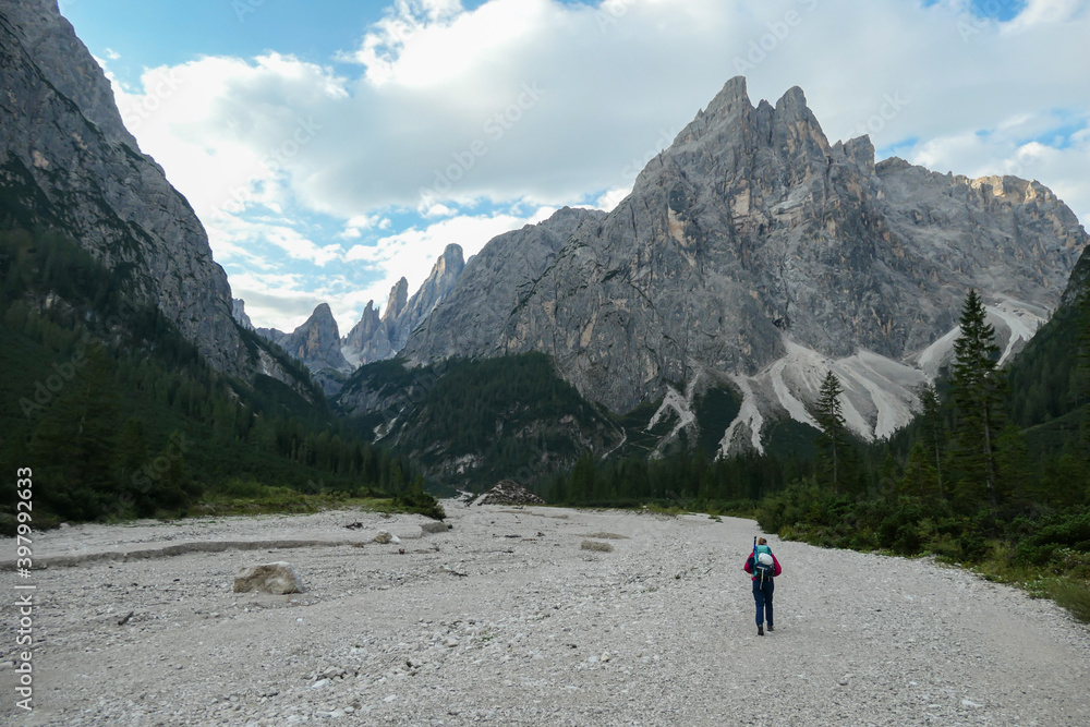 A woman with a big hiking backpack hiking in the Italian Dolomites. There are high and sharp mountain peaks in front of her. She walks on a wide, gravelled road. Experiencing the nature.