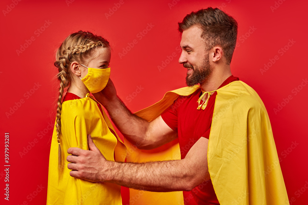 Obraz premium caucasian superhero man is responsible for child girl, he puts a mask on her face , isolated over red background