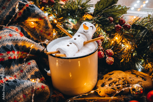 Christmas food white cup with hot chocolate snowman marshmallows. homemade cookie, cinnamon sticks fir xmas tree branches with warm garland lights. New year celebration. Festive dessert breakfast idea photo