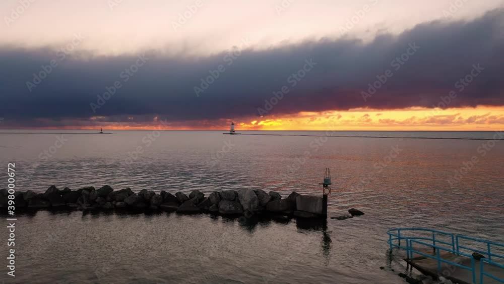 Beautiful sunset aerial flying low over a stone breakwater wall towards the Ludington Breakwater Lighthouse over the water of Lake Michigan with pink and orange sunset clouds above. Beautiful sunset aerial flying low over a stone breakwater wall towards the Ludington Breakwater Lighthouse over the water of Lake Michigan with pink and orange sunset clouds above.