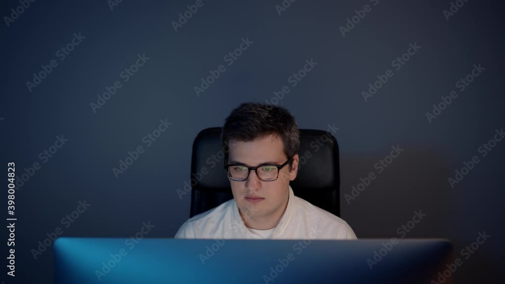 Man in eyeglasses in front of the computer in home office on background of grey wall. Young man sitting at home working alone at night Man in eyeglasses in front of the computer in home office on background of grey wall. Young man sitting at home working alone at night