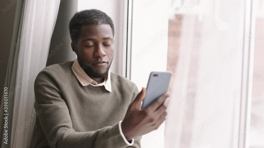 Lockdown of young African man wearing casual clothes sitting on windowsill with mobile phone in his hand and talking to friend by video connection Lockdown of young African man wearing casual clothes sitting on windowsill with mobile phone in his hand and talking to friend by video connection