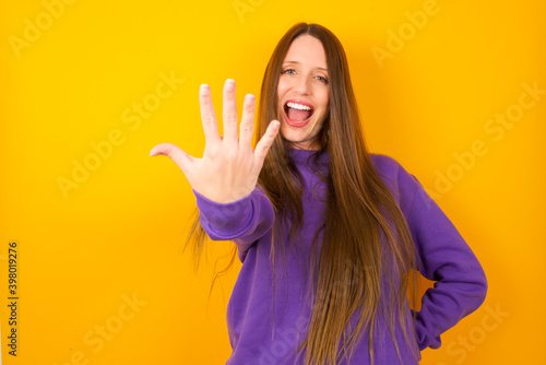 Young beautiful Caucasian woman wearing purple sweater against yellow wall smiling and looking friendly, showing number five or fifth with hand forward, counting down