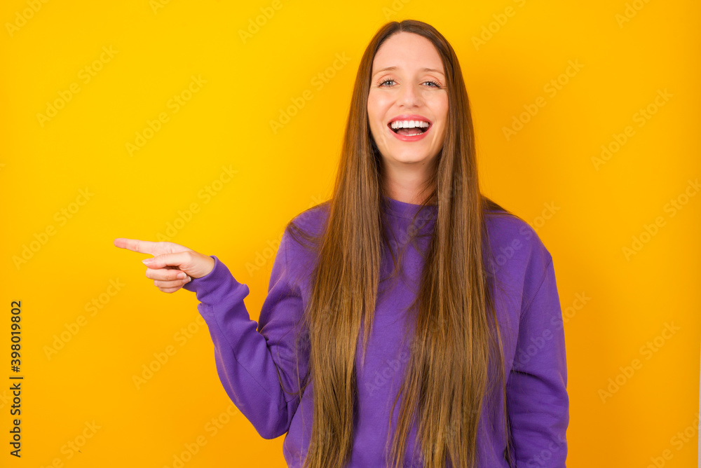 Φωτογραφία Young beautiful Caucasian woman wearing purple sweater against yellow wall laughs happily points away on blank space demonstrates shopping discount offer, excited by good news or unexpected sale