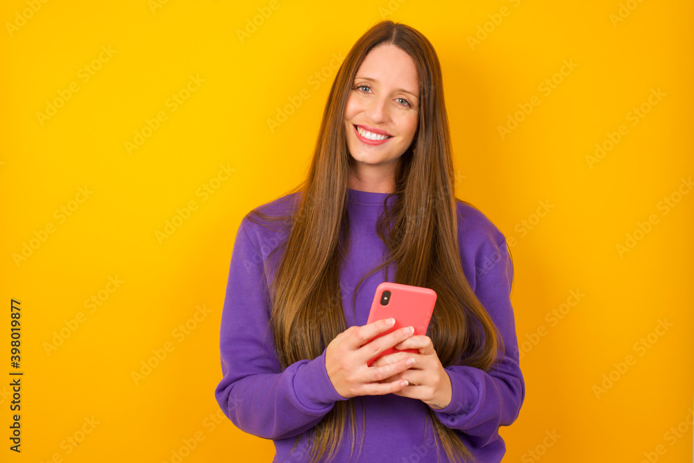Young beautiful Caucasian woman wearing purple sweater against yellow wall Mock up copy space. Using mobile phone, typing sms message Young beautiful Caucasian woman wearing purple sweater against yellow wall Mock up copy space. Using mobile phone, typing sms message