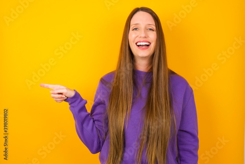 Young beautiful Caucasian woman wearing purple sweater against yellow wall laughs happily points away on blank space demonstrates shopping discount offer, excited by good news or unexpected sale. © Roquillo