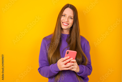 Young beautiful Caucasian woman wearing purple sweater against yellow wall Mock up copy space. Using mobile phone, typing sms message