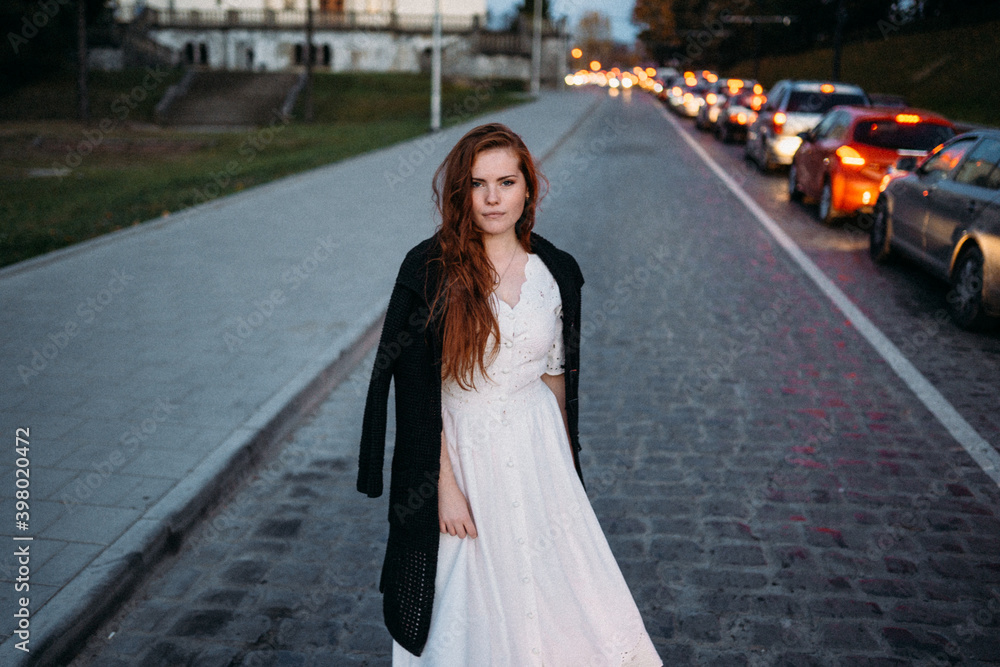 red-haired young woman in a white dress in autumn runs down the street with cars