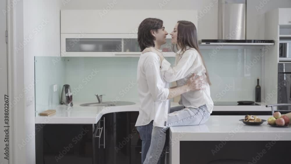 Side view of happy Caucasian young man raising slim beautiful woman on tabletop and kissing adorable girlfriend or wife. Cheerful young couple enjoying weekends at home. Love and leisure concept. Side view of happy Caucasian young man raising slim beautiful woman on tabletop and kissing adorable girlfriend or wife. Cheerful young couple enjoying weekends at home. Love and leisure concept.