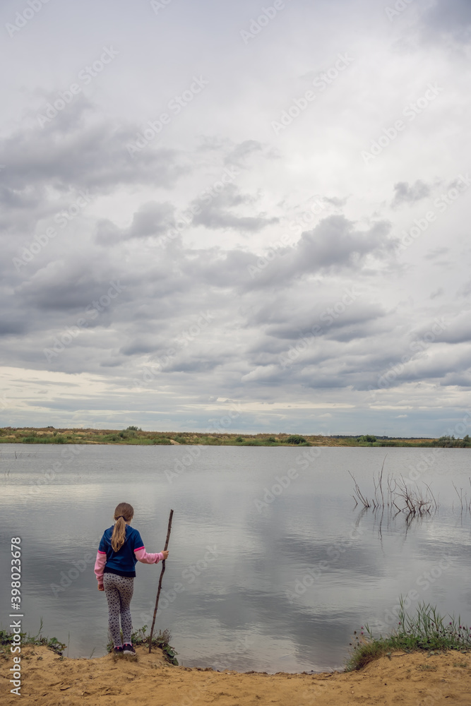 Shallow focus of a kid with a wooden stick for fishing standing near the lake under a cloudy sky Shallow focus of a kid with a wooden stick for fishing standing near the lake under a cloudy sky