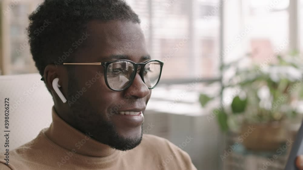 Close up of young African-American male office worker wearing smart casual clothes, glasses and wireless headphones sitting at desktop in office with phone in his hands and having chat Close up of young African-American male office worker wearing smart casual clothes, glasses and wireless headphones sitting at desktop in office with phone in his hands and having chat