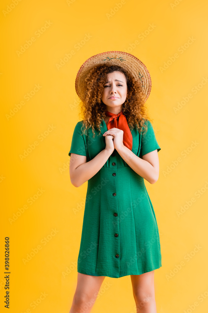 sad and curly woman in straw hat and green dress standing with clenched hands on yellow sad and curly woman in straw hat and green dress standing with clenched hands on yellow