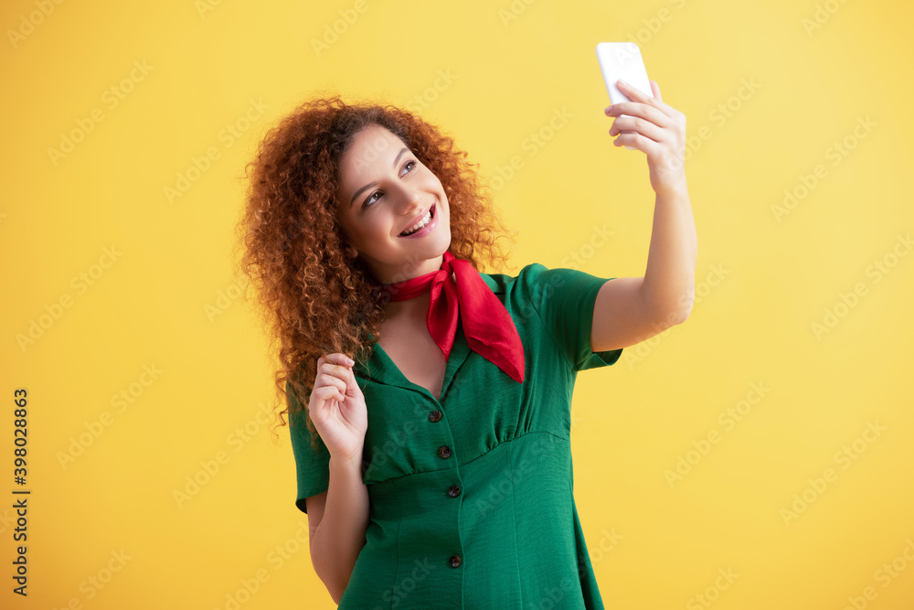 curly young woman in green dress smiling while taking selfie on yellow curly young woman in green dress smiling while taking selfie on yellow