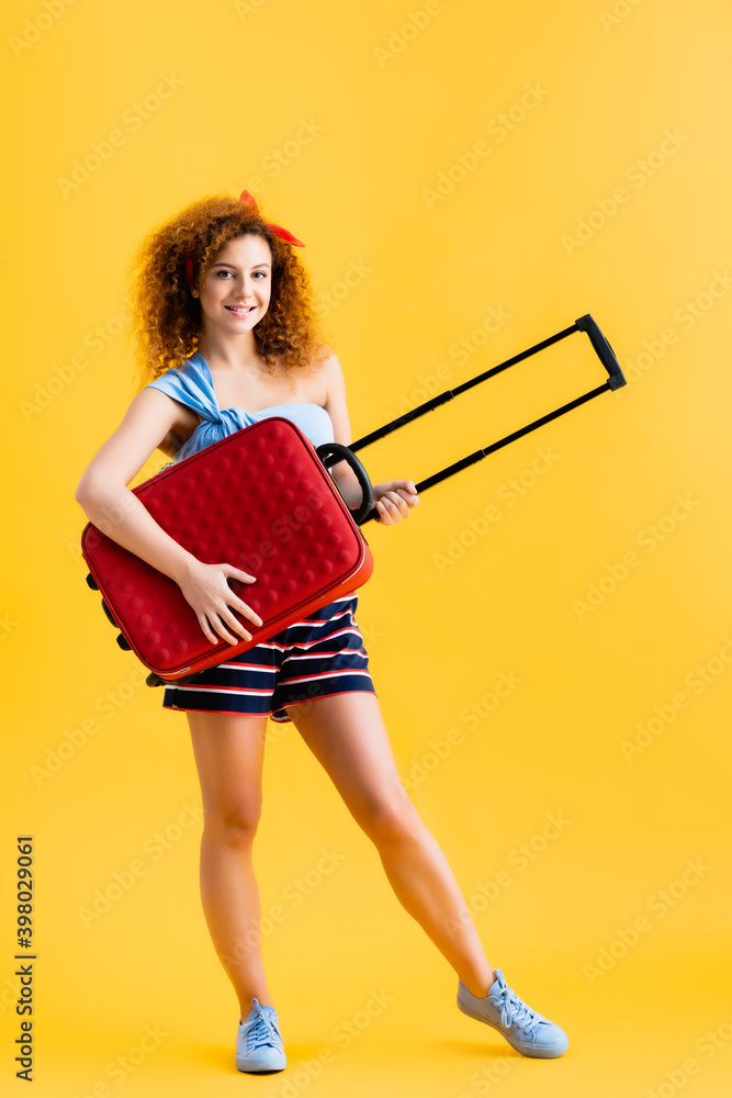full length of happy woman in summer outfit and sneakers holding red suitcase on yellow full length of happy woman in summer outfit and sneakers holding red suitcase on yellow