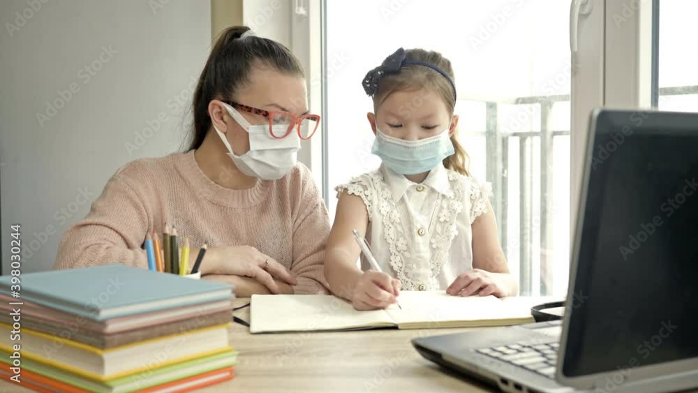 Mom or teacher or tutor helps an elementary school student with her lessons. Both have medical masks. Training during the COVID-19 pandemic. Mom or teacher or tutor helps an elementary school student with her lessons. Both have medical masks. Training during the COVID-19 pandemic.