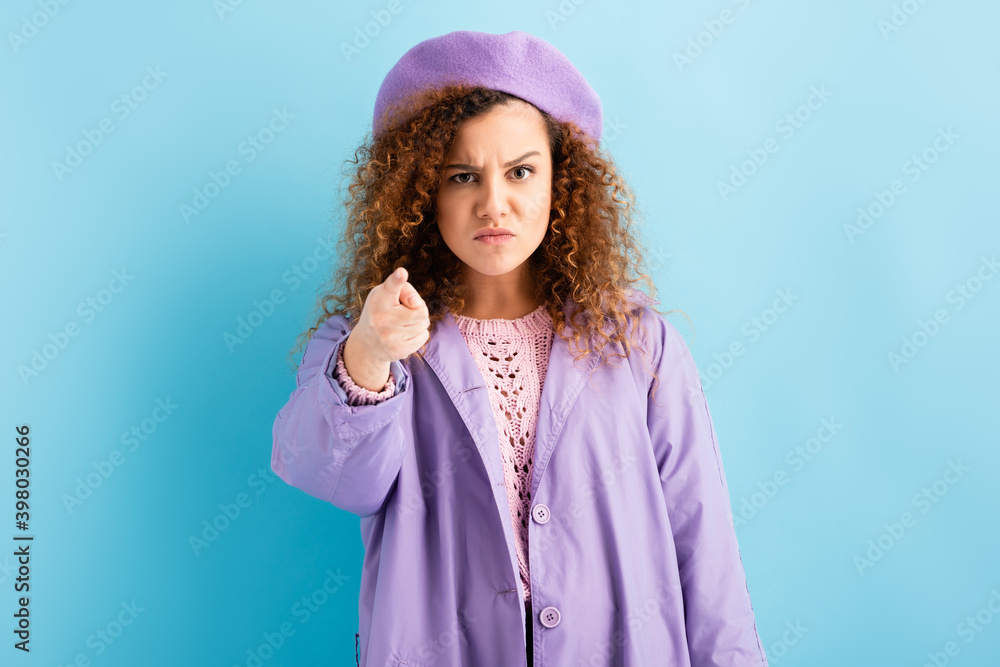angry woman in beret and coat pointing with finger while looking at camera on blue angry woman in beret and coat pointing with finger while looking at camera on blue