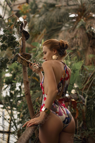 A beautiful girl on swimsuit with snake, natural make-up and red hair stands in the jungle among exotic plant.