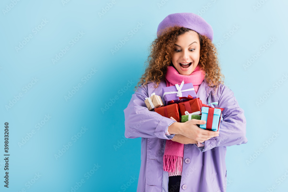 excited woman in winter coat, beret and pink knitted scarf holding christmas presents on blue excited woman in winter coat, beret and pink knitted scarf holding christmas presents on blue