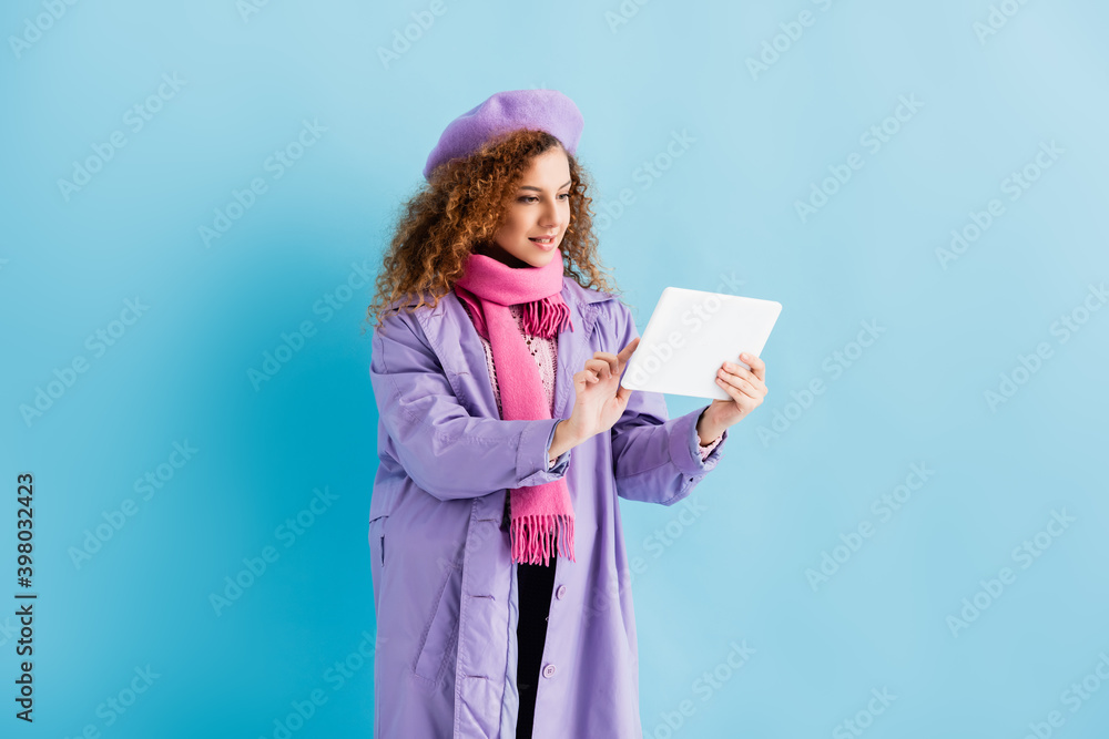 young woman in winter coat, beret and pink knitted scarf using digital tablet on blue young woman in winter coat, beret and pink knitted scarf using digital tablet on blue