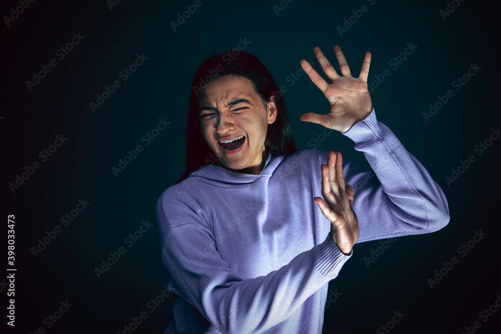 Trying to hide. Portrait of young crazy scared and shocked caucasian woman isolated on dark background. Copyspace for ad. Bright facial expression, human emotions concept. Looking horror on TV, cinema Trying to hide. Portrait of young crazy scared and shocked caucasian woman isolated on dark background. Copyspace for ad. Bright facial expression, human emotions concept. Looking horror on TV, cinema