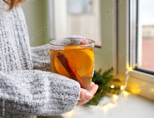 A woman`s hands in a sweater hold a cup of tea with orange and cinnamon by the window. Hot winter flavored drink. Green branches of the Christmas tree