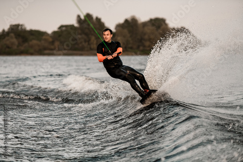 adult man in wetsuit holds cable in his hands and wakeboarding on splashing wave