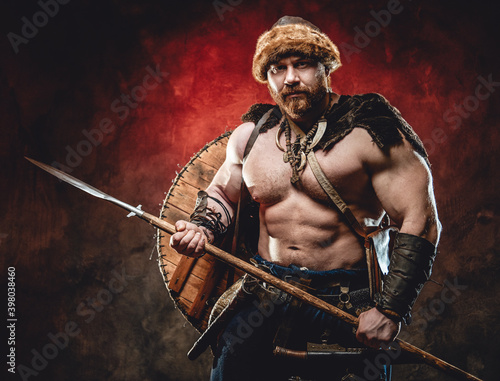 Nude handsome northern barbarian with naked torso and muscular build poses holding a spear in dark red background. © Fxquadro