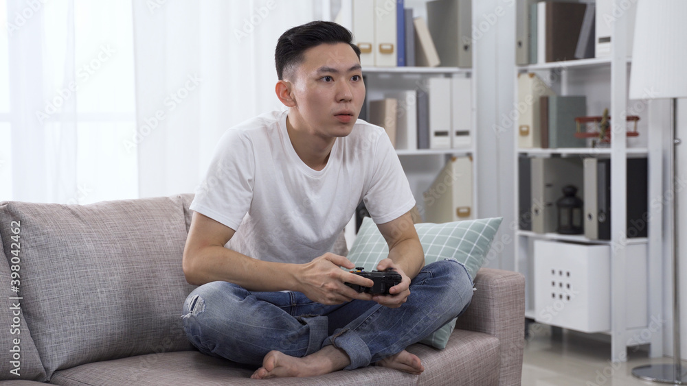 handsome asian man holding wireless controller is playing video game in concentration on sofa in bright living room at home