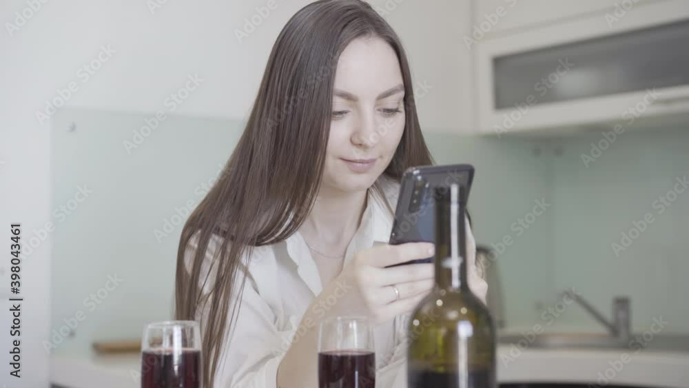 Portrait of absorbed smiling charming woman in kitchen with wine bottle and two glasses messaging online. Portrait of beautiful happy Caucasian girlfriend waiting for boyfriend at home. Dating concept Portrait of absorbed smiling charming woman in kitchen with wine bottle and two glasses messaging online. Portrait of beautiful happy Caucasian girlfriend waiting for boyfriend at home. Dating concept