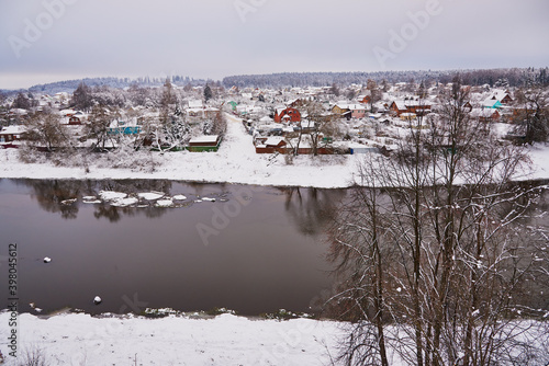 Snow-covered village with colourful wooden houses on the banks of the river, in a cloudy winter day.