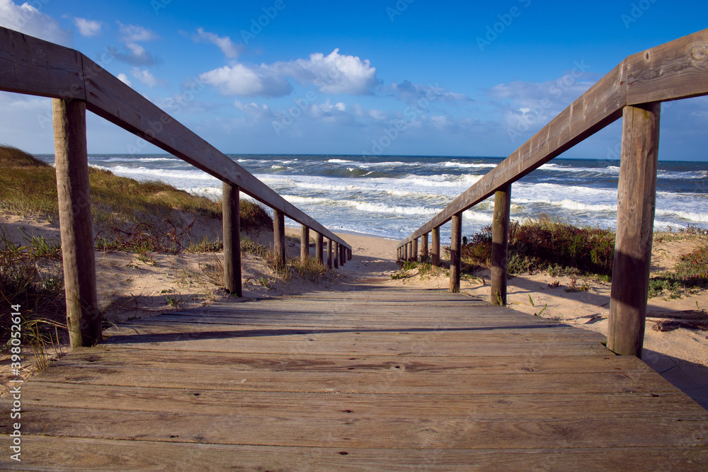 Low angle view of beach wooden boardwalk leading to the rough waves of the Atlantic Ocean. Landscape photography at Quiaios Beach in Portugal