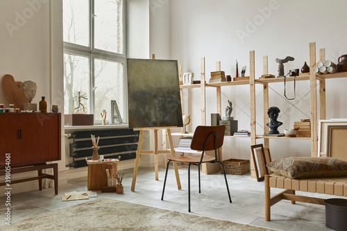 Unique artist workspace interior with stylish teak commode, wooden easel, bookcase, artworks, painting accessories, decoration and elegant personal stuff. Modern work room for artist. Template.