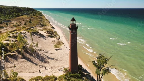 Beautiful aerial view circling around Little Sable Lighthouse along the shoreline of Lake Michigan sitting amongst vegetation and tree covered sand dunes with a clear blue sky above.