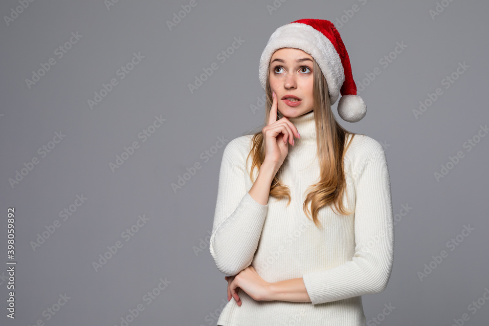 Young beautiful woman wearing a Santa hat over gray background thinking and looking to the side Young beautiful woman wearing a Santa hat over gray background thinking and looking to the side