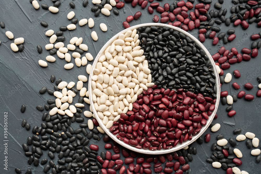 beans in white ceramic plate. bowl of beans. black bean, red kidney bean and white bean. navy bean, cannellini bean, white kidney bean on dark gray background and scattered various beans