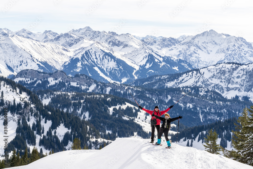 Senior couple is snowshoe hiking in alpine snow winter mountains panorama. Happy successful People with raised arms. Allgau, Bavaria, Germany.
