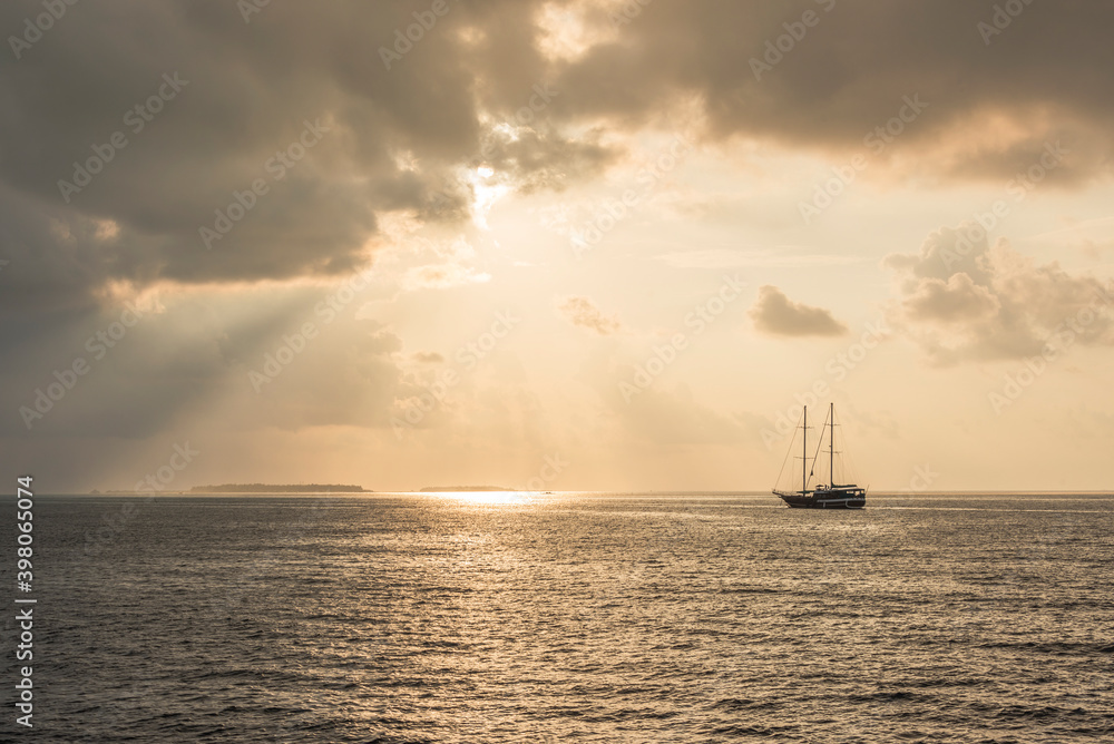 Sun at sunset shining through a picturesque clouds over the Indian ocean with Maldivian islands on the horizon and the silhouette of a ship with masts