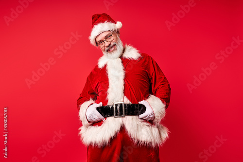 santa claus isolated over red background, studio shoot. senior bad santa with big abdomen is posing at camera and has nasty smile