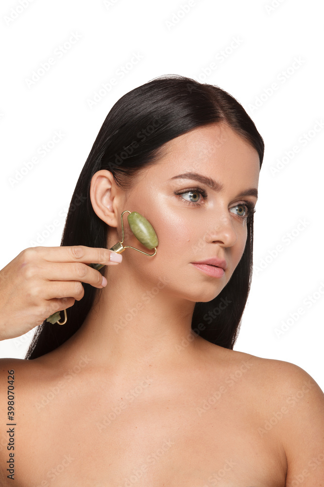 Beautiful young woman with perfect skin wearing towel on head using a jade face roller with natural quartz stones on white background Beautiful young woman with perfect skin wearing towel on head using a jade face roller with natural quartz stones on white background