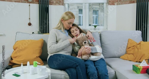 Likable caring adult blond mother hugging her unhappy sick teen daughter while she measuring temperature,holding thermometer in the mouth,close up photo