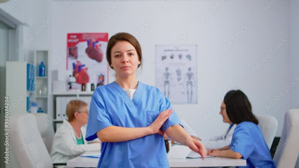 Nurse looking at camera smiling standing in medical conference meeting office. Team of specialist doctors working in background talking about symptoms of disease in clinic room Nurse looking at camera smiling standing in medical conference meeting office. Team of specialist doctors working in background talking about symptoms of disease in clinic room