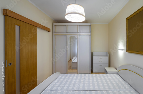 Contemporary interior of bedroom in modern flat. White furniture. Sliding wooden door. Wardrobe with mirror.