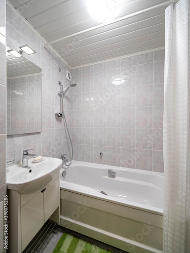 Modern light interior of bathroom in apartment. White tile. Bath tub  sink and mirror. Green mat.