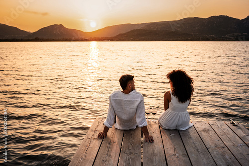 Romantic scene of couple in love at sunset sitting on wooden jetty with wine glasses - Hispanic curly brunette sitting on a pier of lake with orange water reflection looking to her handsome boyfriend