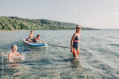 Mom swimming with kids on beach. Caucasian woman mother riding kids children on paddle sup surfboard in lake water. Modern outdoors summer fun family activity. Seasonal recreational sport hobby. © anoushkatoronto