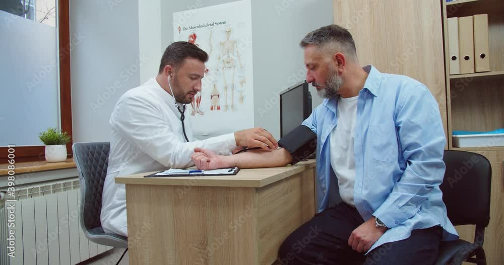 Side view of professional man physician in medical mask sitting in clinic cabinet and measuring blood pressure listening pulse of Caucasian middle-aged patient man with manometer at appointment Side view of professional man physician in medical mask sitting in clinic cabinet and measuring blood pressure listening pulse of Caucasian middle-aged patient man with manometer at appointment