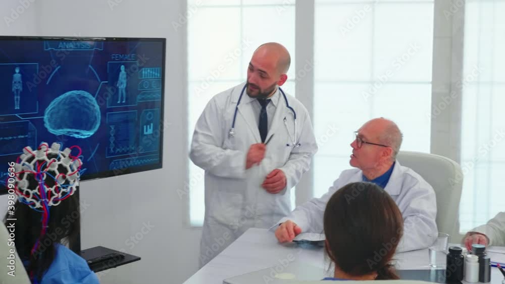 Doctor talking about brain activity during conference with medical staff and nurse wearing headset with sensors. Monitor shows modern brain study while team of scientist adjusts the device. Doctor talking about brain activity during conference with medical staff and nurse wearing headset with sensors. Monitor shows modern brain study while team of scientist adjusts the device.