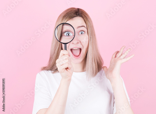 Young attractive blonde woman shocked while looking through a magnifying glass over pink background.